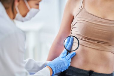 Maskeli bir dermatolog kadın hastasını kontrol ediyor.