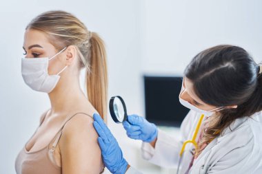 Maskeli bir dermatolog kadın hastasını kontrol ediyor.
