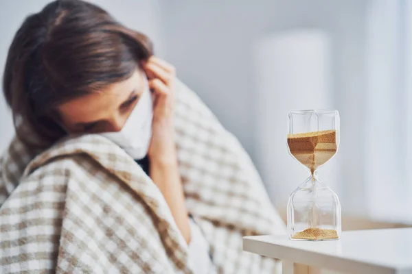 Erwachsene Frau in Maske fühlt sich zu Hause krank mit Sanduhr Covid-19-Konzept — Stockfoto