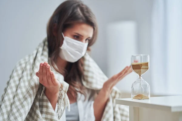 Ενήλικες γυναίκα με μάσκα αισθάνονται άρρωστοι στο σπίτι με hourglass Covid-19 έννοια — Φωτογραφία Αρχείου