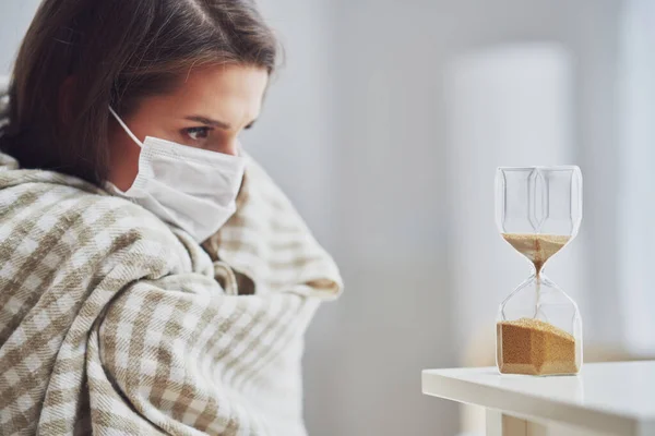 Доросла жінка в масці відчуває себе хворим вдома з пісочним годинником Концепція Covid-19 — стокове фото