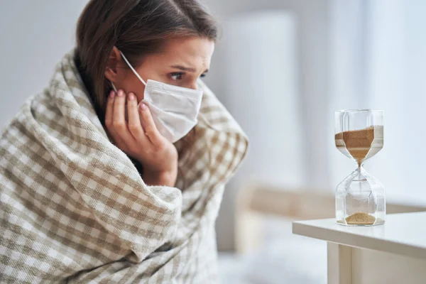 Erwachsene Frau in Maske fühlt sich zu Hause krank mit Sanduhr Covid-19-Konzept — Stockfoto