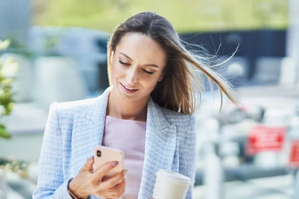 Volwassen aantrekkelijke zakenvrouw met smartphone en koffie wandelen in de stad — Stockfoto