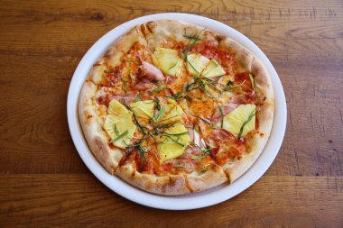 Hawaii pizzası. Öğle yemeği için ahşap bir masada beyaz bir tabakta jambonlu ve ananaslı pizza. Pizza aç insanlar tarafından dünya çapında sevilir. Her yerde pizza.. 