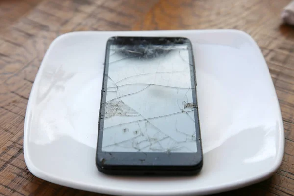 Broken Cellular Telephone. A Cellular Telephone with a cracked and broken Screen on a white plate on a table. Broken Cell Phone.