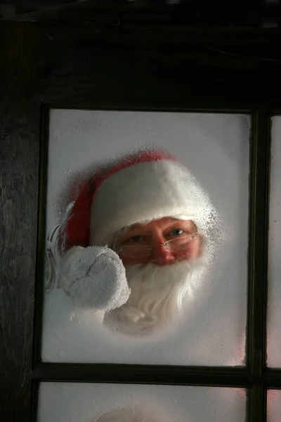 クリスマスだ サンタクロースは雪の窓から外を見る サンタクロースは雪を拭き取り 北極の彼のワークショップウィンドウから霧 メリークリスマス ハッピー ホリデー 季節ごあいさつ サンタクロースはクリスマスの彼の仕事の店で — ストック写真