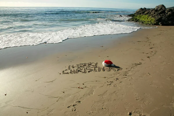 圣诞快乐 节日快乐 写在沙滩上沙滩上的字 圣诞老人的帽子躺在沙滩上 海浪汹涌 沙滩上写着几个字 在海滩的圣诞假期 带着圣诞帽和欢乐假日的海浪 — 图库照片