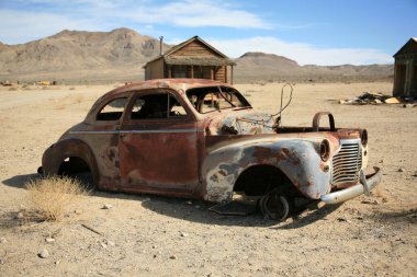 Terk edilmiş araba. Eski bir araba enkazı terk edilmiş. 1800 'lerin California Hayalet Kasabası. Arka planda eski bir altın madeni kasabası var. Kaliforniya Çölü 'nde terkedilmiş bir altın madenciliği kasabasında terkedilmiş bir arabanın arkasında çöken ahşap bir ev.. 