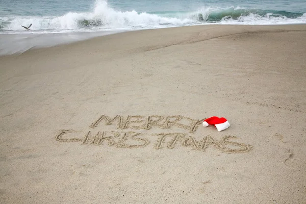 Christmas. Merry Christmas. The words Merry Christmas written in the sand on the beach with a Santa Hat. Words in sand on the beach. Santa\'s hat on a tropical beach. Merry Christmas written in sand with ocean waves and blue sky. Merry Christmas.