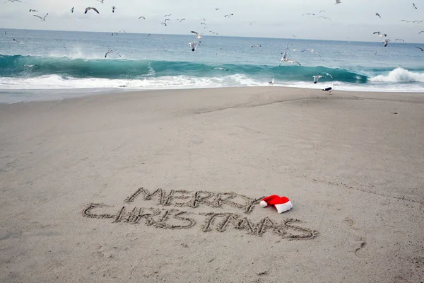 Christmas. Merry Christmas. The words Merry Christmas written in the sand on the beach with a Santa Hat. Words in sand on the beach. Santa's hat on a tropical beach. Merry Christmas written in sand with ocean waves and blue sky. Merry Christmas.