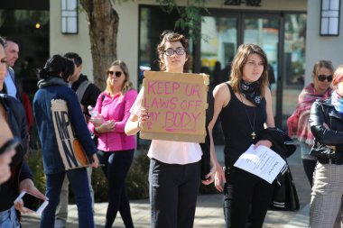 SANTA ANA, CALIFORNIA - JANUARY 21, 2017: People from Orange County carry signs, and wear t-shirts with slogans marched in Santa Ana today for the Orange County Women's March. President Donald J. Trump Protesters. Pink Pussy Hats. Protest Signs.  clipart