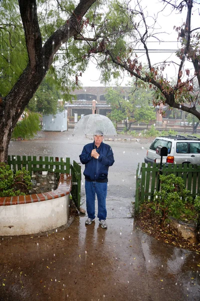 Rain Storm. Rain. Rain drops rippling in a puddle. Rain drops and splashing of pouring rain. A man stands outside in the pouring rain wearing a blue rain coat and a clear umbrella. A hose attached to a house rain gutter to divert rain water away.