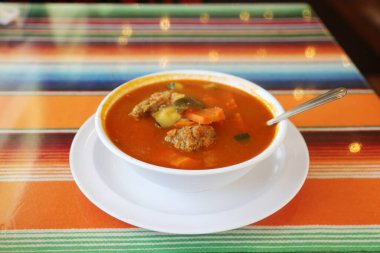 Meksika yemeği. Meksika Lokantası. ALBONDIGAS SOUP. Cips ve salsa sosu. Meksika yemeği Meksika çorbası köfteli ve sebzeli. Meksika albondiga çorbası, beyaz bir seramik kâse ve tabak, geleneksel Meksika serapı, boş masa örtüsü ve kaşık..