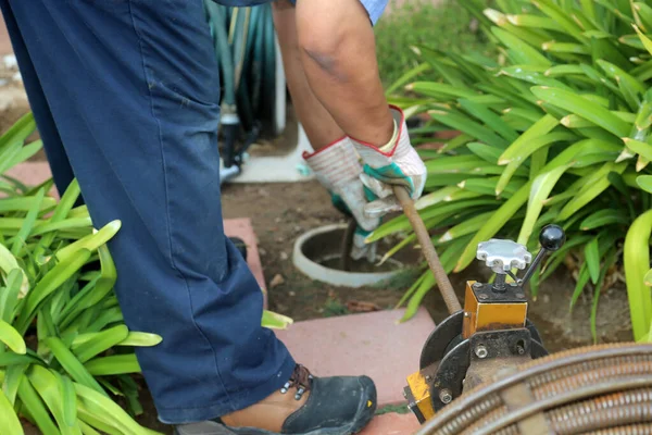 Sewer cleaning. A plumber uses a sewer snake to clean blockage in a sewer line. Plumber holding a drain pipe, providing sewer cleaning service outdoor. Sewage pumping machine is unclogging blocked drain.