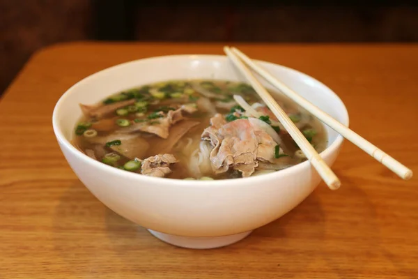 Vietnamese rice noodle soup with sliced rare beef.  Vietnamese Cuisine - Beef Pho Noodle Soup or Pho Bo Soup. Served with Fresh Greens in White Bowl on Wooden Table. Pho Bo Soup with Spicy Sauces. Soup for Lunch. Soup for Dinner. Soup for a Snack.