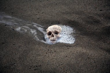 Cadılar Bayramı. İNSAN SKULL. İnsan Kafatası sahile vuruyor. İnsan Kafatası kuma gömüldü. Kumda insan kafatası, dalgalar ve deniz köpüğüyle okyanus. Antik arkeolojik alandaki korkutucu insan kemikleri. Kafatası okyanustaki kumda gömülü..