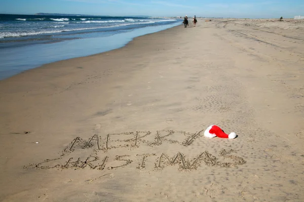Merry Christmas. Words written in sand in San Diego California close to the US/Mexican border. Red and White Santa Claus Hat sits on the sand next to the text.