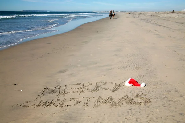 Merry Christmas. Words written in sand in San Diego California close to the US/Mexican border. Red and White Santa Claus Hat sits on the sand next to the text.