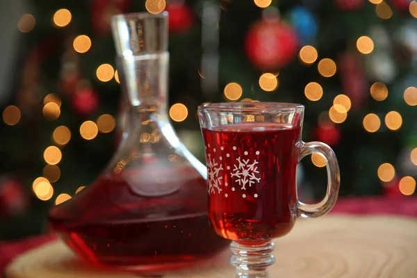 Christmas Wine. Wine or Juice in a Clear Glass Decanter and festive glass cup. Christmas Tree out of focus in background. Room for text Overlay. Christmas Images. Tree Trunk Cutting board on red.