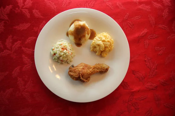 Chicken Dinner. Close Up View of a Chicken Dinner. White plate. Red Table Cloth. Chicken Leg. Biscuit. Coleslaw. Mashed Potatoes and Gravy. Macaroni and Cheese. A Meal fit for a King.