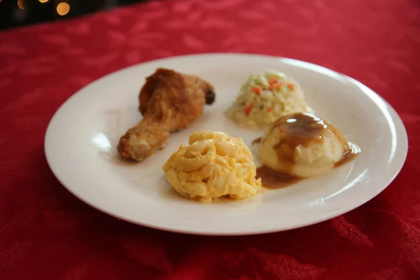 Chicken Dinner. Close Up View of a Chicken Dinner. White plate. Red Table Cloth. Chicken Leg. Biscuit. Coleslaw. Mashed Potatoes and Gravy. Macaroni and Cheese. A Meal fit for a King.