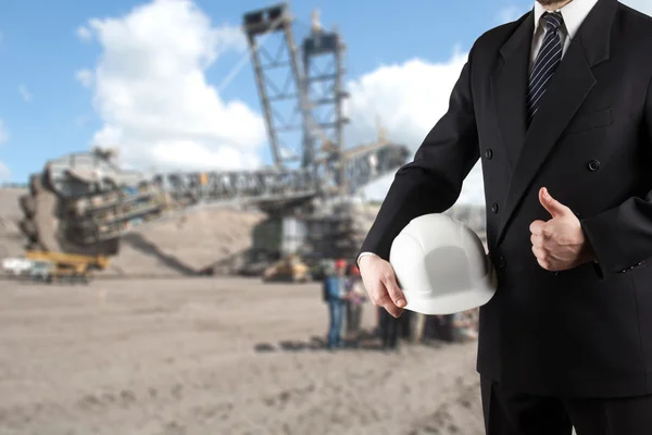 Primer plano de la mano del ingeniero sosteniendo el casco de seguridad blanco para la seguridad de los trabajadores de pie frente a la obra borrosa con grúas en el fondo — Foto de Stock