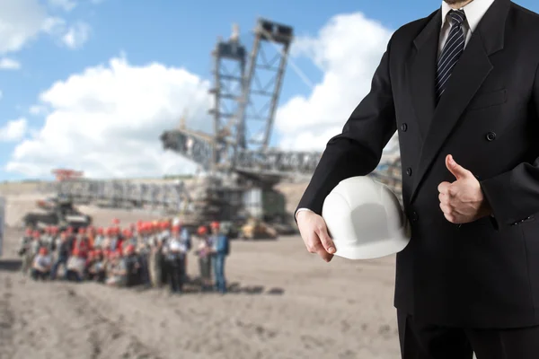 Primer plano de la mano del ingeniero sosteniendo el casco de seguridad blanco para la seguridad de los trabajadores de pie frente a la obra borrosa con grúas en el fondo — Foto de Stock