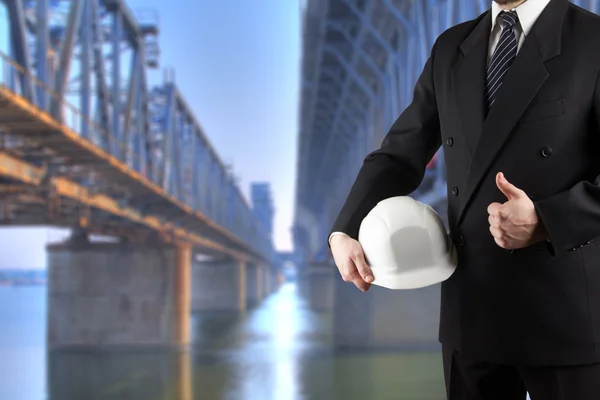 Primer plano de la mano del ingeniero sosteniendo el casco de seguridad blanco para la seguridad de los trabajadores de pie frente a la obra borrosa con grúas en el fondo — Foto de Stock