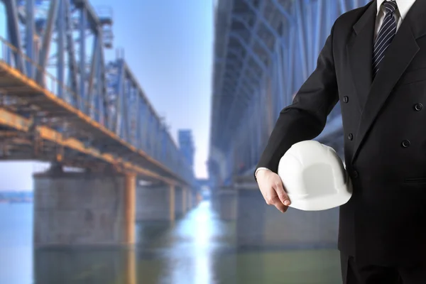 Primer plano de la mano del ingeniero sosteniendo el casco de seguridad blanco para la seguridad de los trabajadores de pie frente a la obra borrosa con grúas en el fondo — Foto de Stock