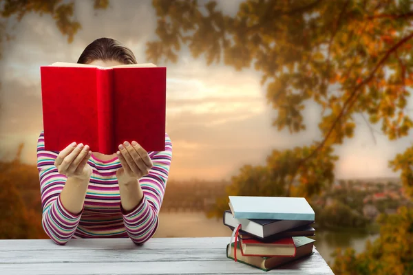 Young woman reading a book and covering her face ,sitting by wooden table with stack of colorful hardback books on blurred nature landscape backdrop.