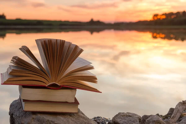 Stack of books and Open hardback book on blurred nature landscape backdrop against sunset sky with light. Copy space, back to school. Education background. — Stock Photo, Image