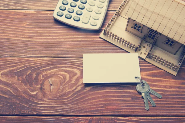 Concepto de Bienes Raíces. Casa modelo, llaves, tarjeta de visita en blanco y calculadora en mesa de madera. Vista superior. Imagen tonificada — Foto de Stock