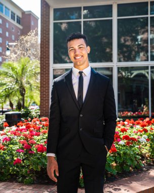 Genç, kendine güvenen, mutlu, gülümseyen bir iş adamı ya da üniversite öğrencisi, önünde duran tuğla binaya kameraya bakıyor.