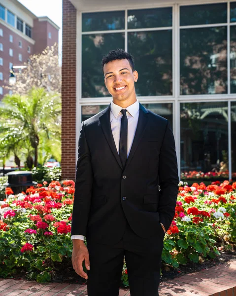 Genç, kendine güvenen, mutlu, gülümseyen bir iş adamı ya da üniversite öğrencisi, önünde duran tuğla binaya kameraya bakıyor.