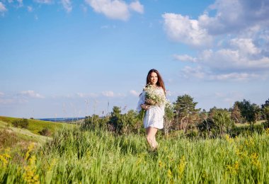 Bir buket çiçekli beyaz elbiseli bir kız tarlada yürüyor.