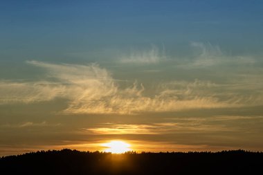 Parlak günbatımı manzaranın üzerine altın renkler saçar. İnce bulutlar ve ön planda koyu ağaç siluetleri olan sakin bir atmosfer yaratır..