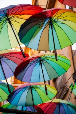 Canlı bir caddenin üzerinde çeşitli renkli şemsiyeler asılı, neşeli gölgeler ve güneşli bir günde açık hava alışverişine cazibe katıyor..