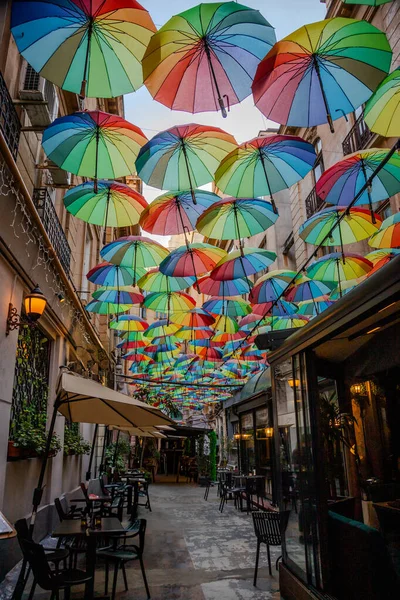 Dar bir şehir sokağı, yukarıdan sarkan parlak renkli şemsiyelerle dekore edilir ve insanlar aşağıdaki kafede içkinin keyfini çıkarırken neşeli bir ortam yaratır..