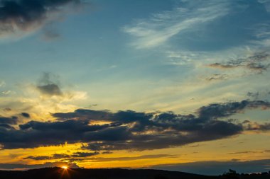 Sersemletici bir günbatımı, turuncu ve mavi tonlarıyla gökyüzünü boyar. Bulutlar, yuvarlanan tepelerin üzerinde tembelce sürüklenirken kırsal alanda sakin bir manzara yaratır..