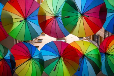 Bir dizi parlak renkli şemsiye dar bir sokakta asılı, açık mavi bir gökyüzü altında şehir ayarı cazibe ve neşe katar.