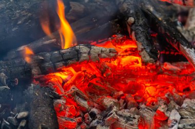 Parlak turuncu alevler kömürleşmiş odun ve küllerle çevrili parlayan közlerden yükseliyor ve açık havada toplanmak için sıcak ve davetkar bir atmosfer yaratıyor..