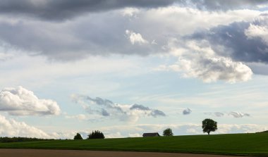 Huzurlu bir kırsal alan, dramatik bulutlarla dolu uçsuz bucaksız gökyüzünün altında uzanan canlı yeşil alanlar barındırır. Yalnız bir ağaç ve uzak yapılar manzaraya derinlik katar..