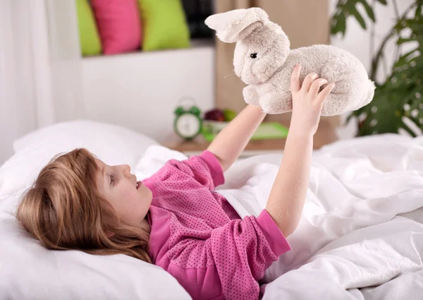 Linda niña rubia acostada en la cama jugando con conejito de peluche —  Fotos de Stock