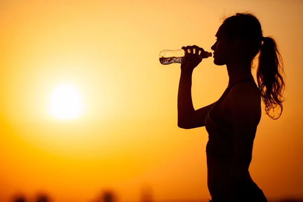 Silueta de mujer joven bebe agua contra el fondo del atardecer — Foto de Stock