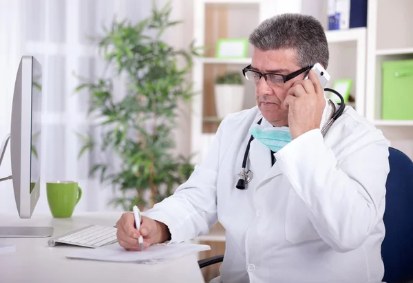 Senior doctor in the office talking on a mobile phone and writes — Stock Photo, Image