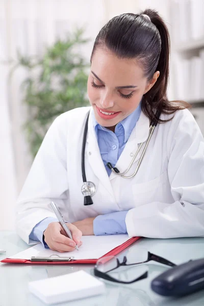 Schöne junge lächelnde Ärztin sitzt am Schreibtisch und wr — Stockfoto