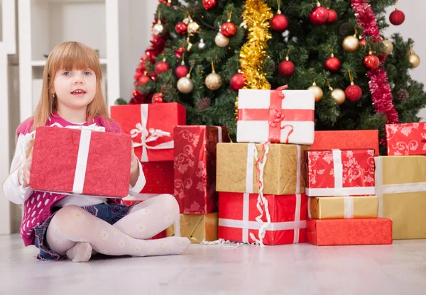 Csinos kislány mosolyog ajándékkal a karácsonyfa mellett — Stock Fotó