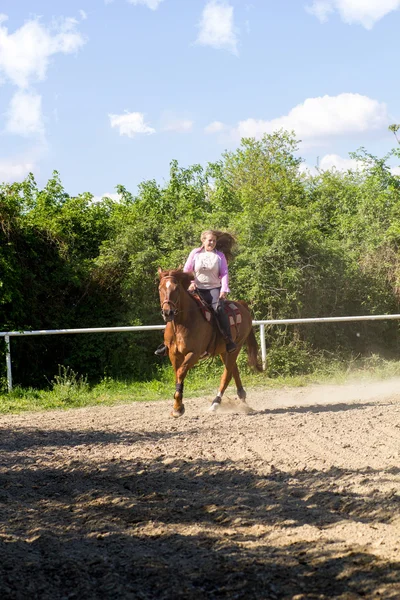 Jovem sorrindo menina montando seu cavalo marrom em um campo de treinamento — Fotografia de Stock