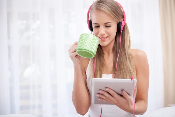 Unga blonda sittande på sängen lyssnar på musik och dricka te — Stockfoto