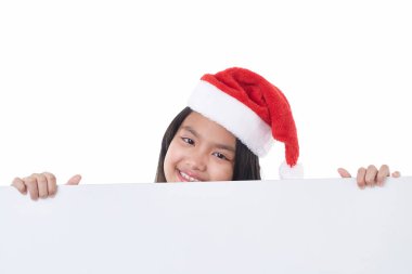 Noel Baba şapkası takan mutlu küçük bir kızın portresi beyaz arka planda izole edilmiş beyaz bir panonun arkasında boş bir Noel panosu tutuyor.